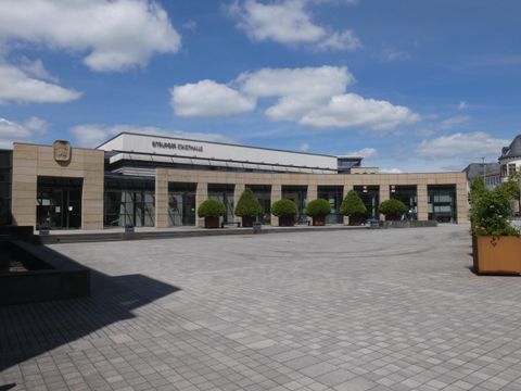 Ein modernes Gebäude mit großen Fenstern und einem klaren Himmel. Der Platz vor dem Gebäude ist gepflastert und mit grünen Sträuchern geschmückt.