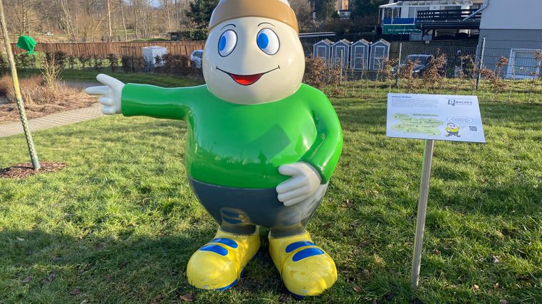 Statue von Maskottchen Willi Basalt mit grünem Shirt, grauer Hose und gelben Schuhen zeigt nach links. Hinter ihm Gebäude und ein Park.