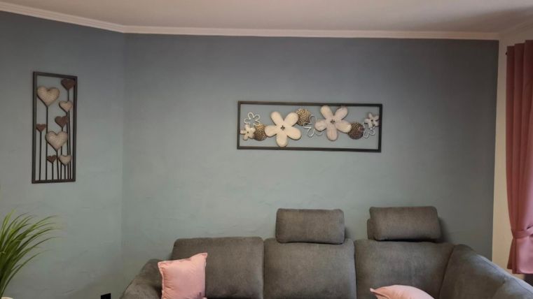 Ein gemütliches Wohnzimmer mit einem grauen Sofa und einer Pflanze. An der Wand hängen dekorative Bilder mit floralen Motiven.