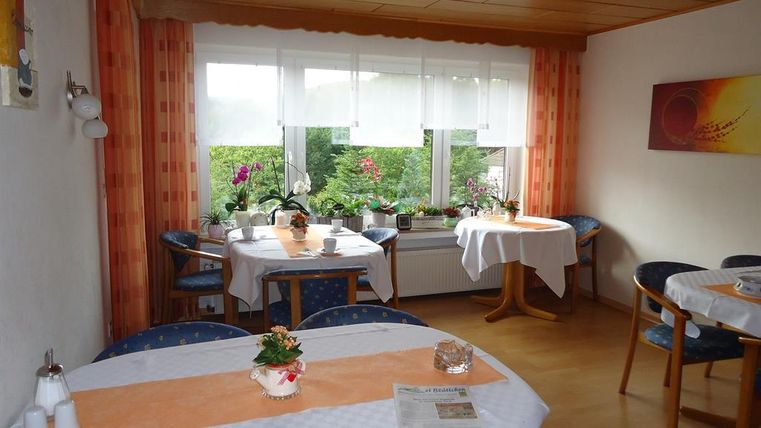 A cozy dining area with tables and chairs. Large windows offer a view of a green garden.