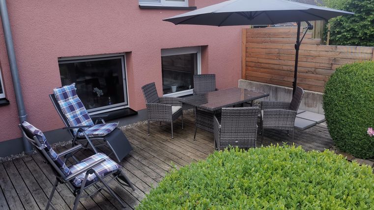 Eine gemütliche Terrasse mit einem großen Tisch und Stühlen unter einem Sonnenschirm. Umgeben von einem grünen Busch und einem Holzzaun.