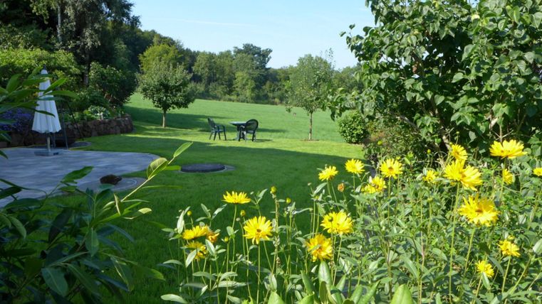 Ein schöner Garten mit leuchtenden gelben Blumen und grünem Rasen. Im Hintergrund stehen einige Stühle und Bäume unter einem klaren Himmel.