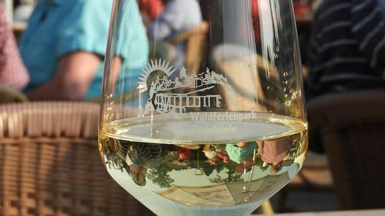 Ein Weinglas mit Gravur "Waldferienpark Gerolstein" steht auf einem Tisch. Im Hintergrund sind unscharf Menschen zu sehen.