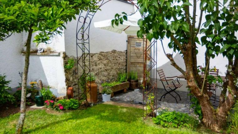 Ein einladender Garten mit grüner Rasenfläche und bunten Pflanzen. Im Hintergrund steht ein Tisch mit Stühlen unter einem schattigen Baum.