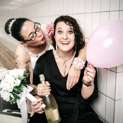 Zwei Frauen in schicker Kleidung posieren zusammen mit einer Flasche Sekt, Luftballon und einem Blumenstrauß in den Händen.