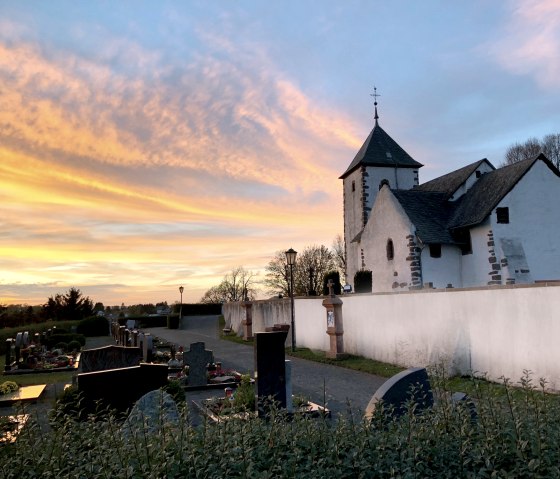Eine Wehrkirche bei Sonnenuntergang, umgeben von einem Friedhof. Der Himmel ist in warmen Orange- und Blautönen gefärbt.