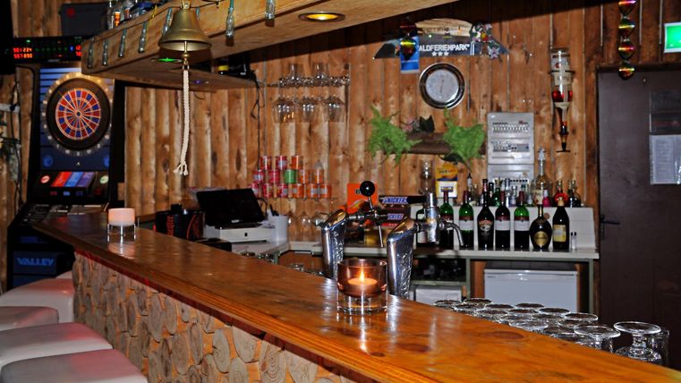 Rustikale Kellerbar mit Holztresen, Zapfhahn, Flaschen, Gläsern und einem Dartautomaten im Hintergrund. Eine Glocke hängt über der Theke.
