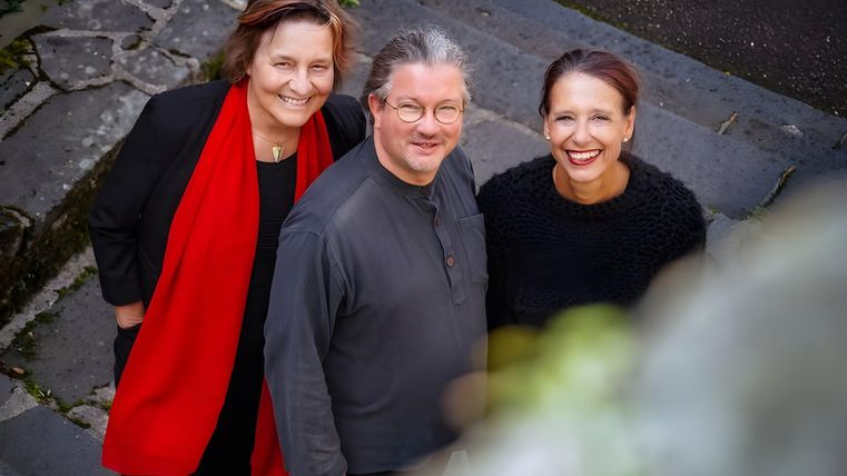 Trois personnes se tiennent souriant sur un escalier. Elles portent différents styles de vêtements et semblent amicales et détendues.