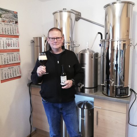 Ein Mann steht vor einer Brauanlage und h&auml;lt eine Flasche Bier und ein gef&uuml;lltes Glas hoch. 