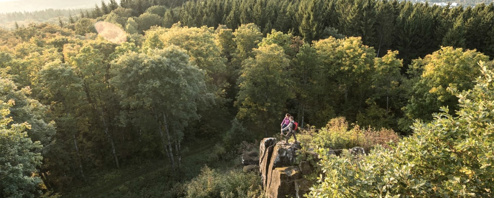 Two people are standing on a rock surrounded by a dense forest. Hills and a wide view over the landscape can be seen in the background., &copy; Eifel Tourismus GmbH, D. Ketz
