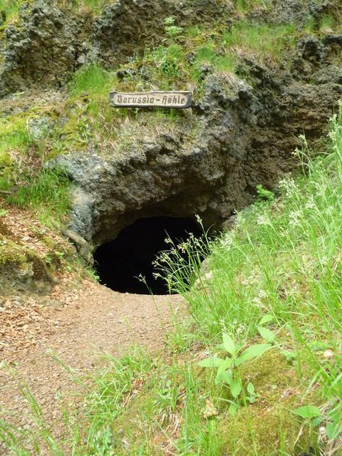 <p>Höhleneingang in einer bewachsenen Felswand. Ein Waldpfad führt zur Höhle und über dem Eingang hängt ein Holzschild.</p>
