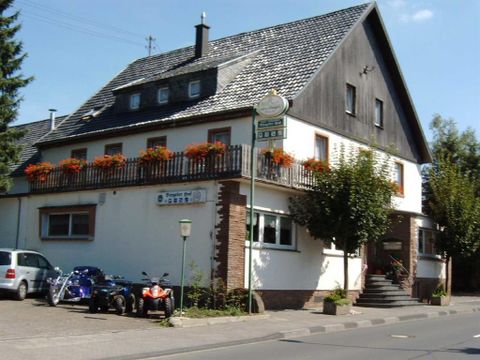 <p>Ein dreigeschossiges Gebäude mit schwarzem Dach.Den Eingang erreicht man über eine Treppe. Im ersten Stock umrahmt ein Holzgeländer mit Blumenkästen einen großen Balkon. Links neben dem Haus ist ein Parkplatz, der mit drei Autos belegt ist. Hinter dem Parkplatz hängt ein Schild, mit der Aufschrift "Biergarten". Vor dem Hotel verläuft ein Bürgersteig, der direkt an die Hauptstrasse angrenzt.</p>