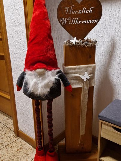 Een decoratieve kerstfiguur in de vorm van een elf met een rode hoed staat naast een welkomstbord van hout. De elf heeft lange benen en draagt een grijze romp met witte vacht.