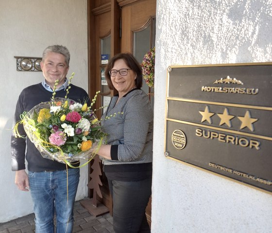 Zwei Personen halten einen Blumestrauß vor der Eingangstür des Landhauses.