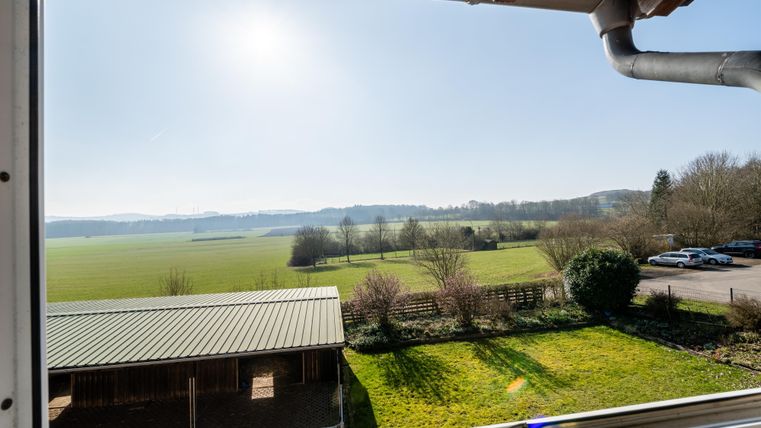 Eine weitläufige Landschaft mit grünen Feldern und sanften Hügeln. Im Vordergrund ist eine Struktur mit einem Metaldach und Bäumen zu sehen.