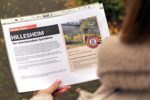Person liest ein Blatt über einen Krimi-Trail in Hillesheim, Titel: Der verschwundene Sportlehrer. Im Hintergrund herbstliche Umgebung.