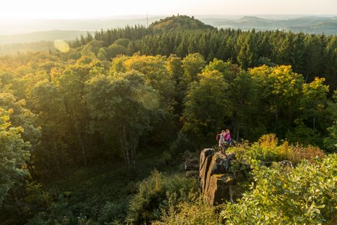 Zwei Personen stehen auf einem Felsen mit Blick auf einen bewaldeten Hügel im Sonnenuntergang.