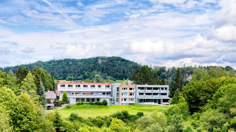Ein modernes Hotelgebäude inmitten grüner Landschaft mit bewaldeten Hügeln im Hintergrund.