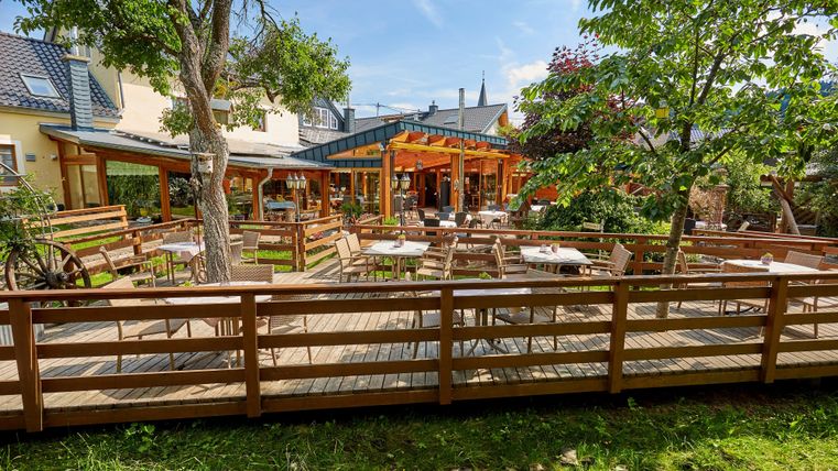 Eine sonnige Terrasse mit Holzmöbeln und Bäumen, umgeben von einem Holzzaun, vor einem Gebäude mit großen Fenstern.