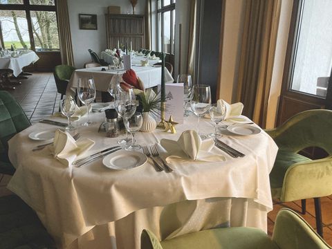 Im Vordergrund ein gedeckter Tisch mit gefalteten Servietten, Weingläsern und Besteck ; Im Hintergrund sieht man den Rest des Raumes mit Tischen