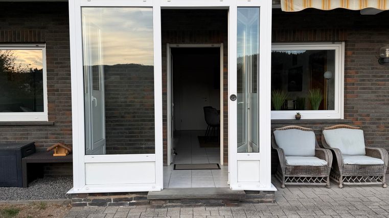 Une entrée accueillante avec de grandes fenêtres et une porte moderne. Sur la véranda, il y a des chaises confortables et l'espace paraît lumineux et amical.