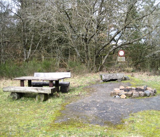 A wooden table with benches stands next to a fireplace in the forest. Trees and a sign can be seen in the background., © Touristik GmbH Gerolsteiner Land, Ute Klinkhammer