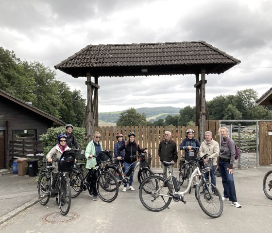 Neun sportlich gekleidete Personen stehen vor dem Eingang des Adler- und Wolfspark Kasselburg und einige Fahrr&auml;dern parken bei ihnen.