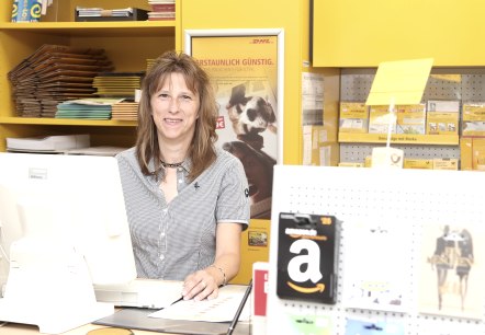 A woman stands at a post office counter and smiles at the camera. In front of her is a screen with a keyboard, and behind her are shelves full of envelopes, parcels and other postal items.