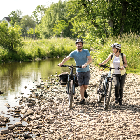 A man and a woman push their bikes over stones on the banks of the river in Stadtkyll spa gardens.