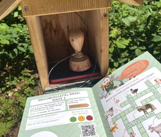 Een wandelpas wordt gestempeld bij een houten stempelstation buiten, omgeven door groene vegetatie., © Touristik GmbH Gerolsteiner Land, Leonie Post