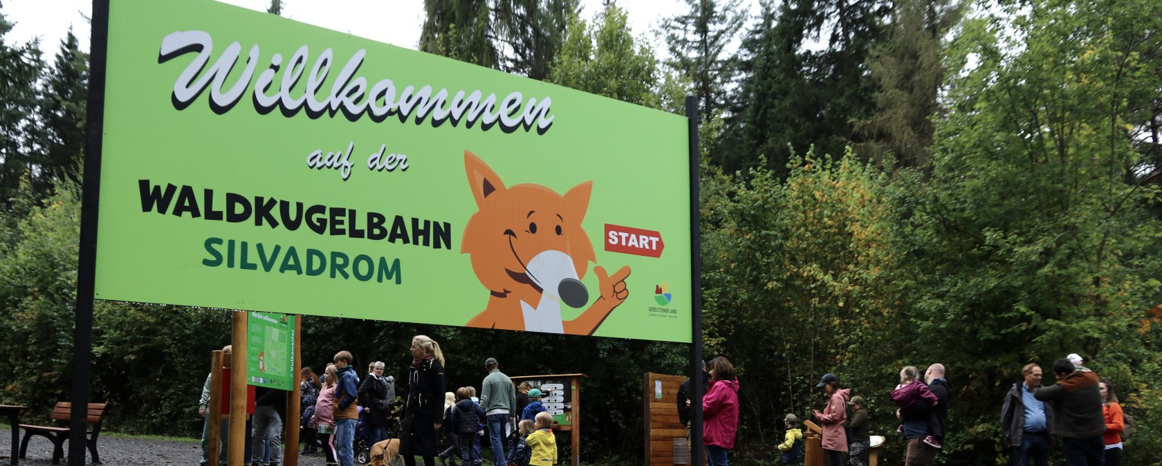 Gro&szlig;es buntes Schild mit Aufschrift "Willkommen auf der Waldkugelbahn Silvadrom" mit Fuchsmaskottchen, der zum Start zeigt. Dahinter laufen zahlreiche Personen.