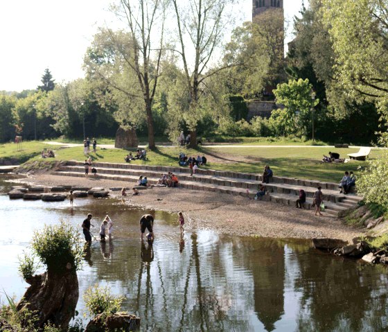 Mensen ontspannen bij het water in Kyllpark, terwijl op de achtergrond een kerktoren te zien is. Kinderen spelen in het ondiepe water., © Clara Zins-Grohé