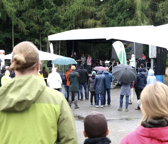 Numerous people wearing rain jackets and carrying umbrellas stand in front of a stage where three people are speaking and musicians are seated.