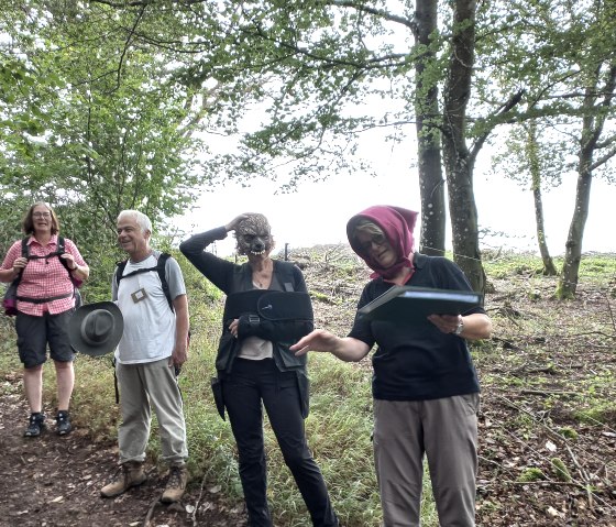 Vier Personen stehen nebeneinder auf einem Waldweg. Die zwei Frauen vorne tragen Verkleidungen und spielen eine Geschichte vor.
