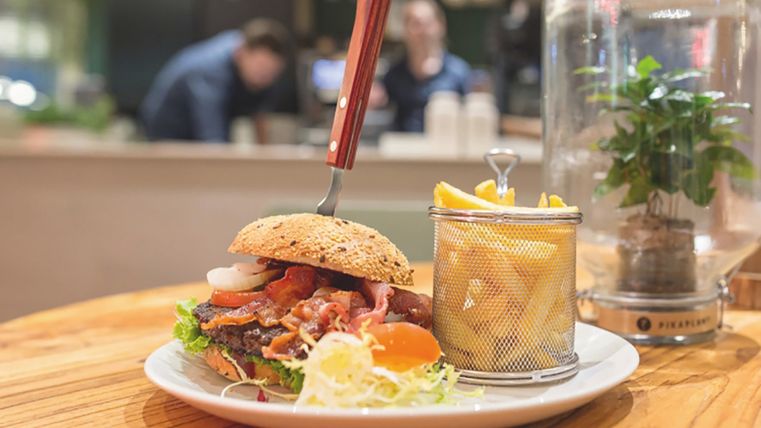 Auf einem Holztisch steht ein Teller mit einem Burger und Pommes darauf, in dem Burger steckt ein Messer, im Hintergrund sieht man arbeitende Menschen