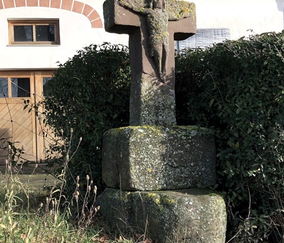 Ein verwittertes Steinkreuz mit Christusfigur steht vor einem Haus mit rotem Ziegeldach und Holztür, umgeben von grüner Vegetation., © Touristik GmbH Gerolsteiner Land