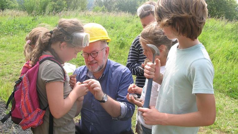 Kinder und ein Erwachsener mit Schutzbrillen und Hammer untersuchen Steine auf einer Wiese.