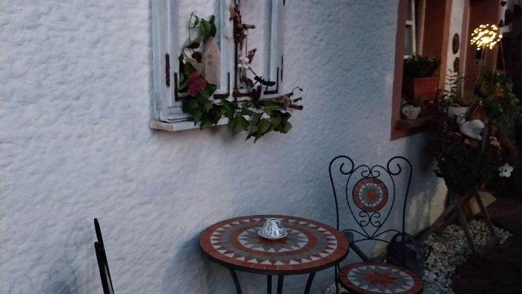 Een gezellige buitenruimte met een ronde tafel en twee metalen stoelen. De wand is wit en een decoratief raam met bloemen geeft de omgeving charme.