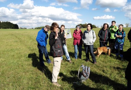 A group of about 9 people stand in a large meadow and look at a woman. The lady holds documents in her hand and explains something to the group.