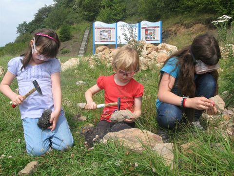 Drei Kinder mit Schutzbrillen und Hämmern untersuchen Steine auf einer Wiese. Im Hintergrund sind Infotafeln zu sehen.