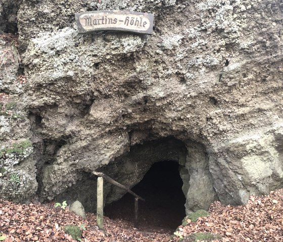 Entrance to Martinshöhle cave in Hohenfels Essingen, surrounded by foliage and rocks. A wooden sign with the inscription 'Martins-höhle' is visible., © Touristik GmbH Gerolsteiner Land
