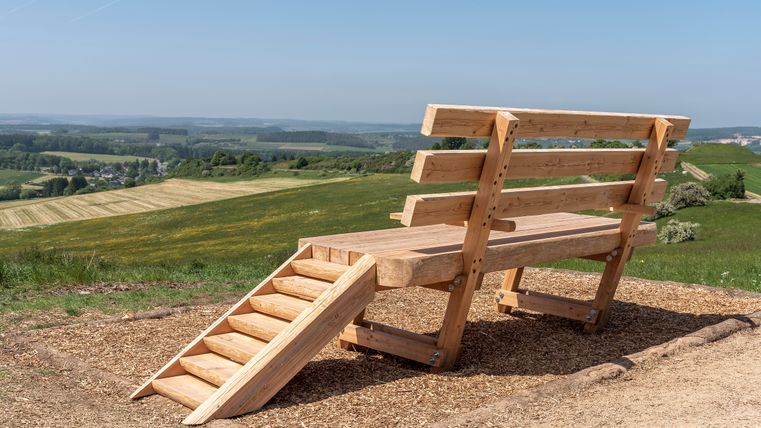 Eine übergroße Holzbank mit Treppe steht auf einem Hügel mit Blick auf Felder und Wälder unter blauem Himmel.