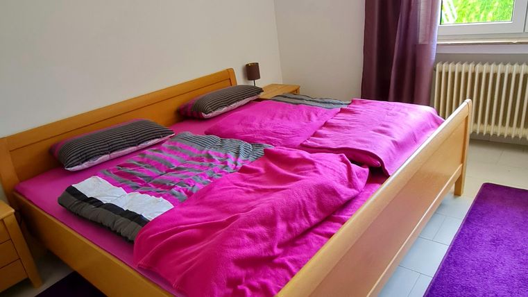 A modern bedroom with a large wooden bed and colorful blankets. The room has a purple carpet and a window with curtains.