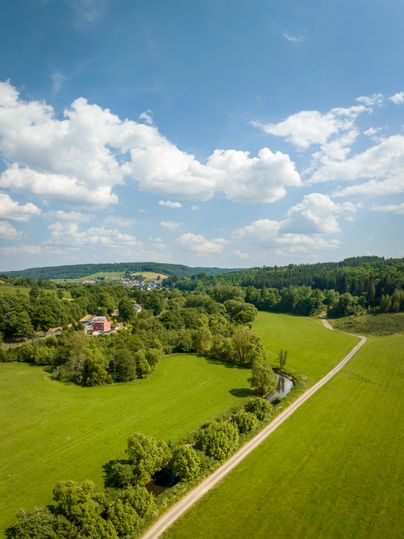 Blick auf eine hügelige Landschaft mit Bäumen und einer Brücke unter einem wolkigen, blauen Himmel