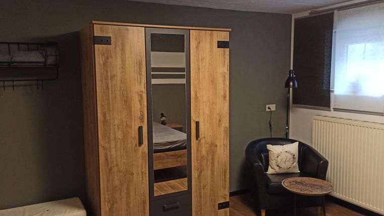 Une chambre simple avec une armoire, un fauteuil et une table. Il y a un lit et une fenêtre qui laisse entrer la lumière naturelle.