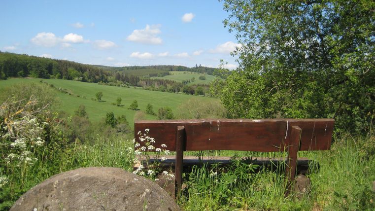 Holzbank auf einem Hügel mit Blick auf grüne Felder und Wälder, blauer Himmel mit wenigen Wolken.
