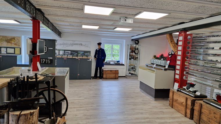 Un musée avec une exposition historique. En arrière-plan, une figure en uniforme se tient tandis que divers objets et vitrines sont dispersés dans la pièce.