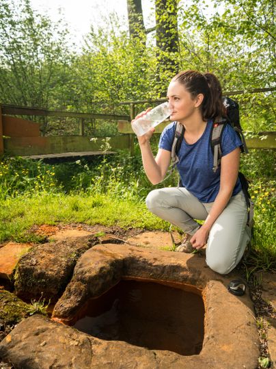 Frau kniet neben einer Quelle im Wald, trinkt aus einer Flasche. Sie trägt Outdoor-Kleidung und einen Rucksack. Umgeben von grüner Vegetation.