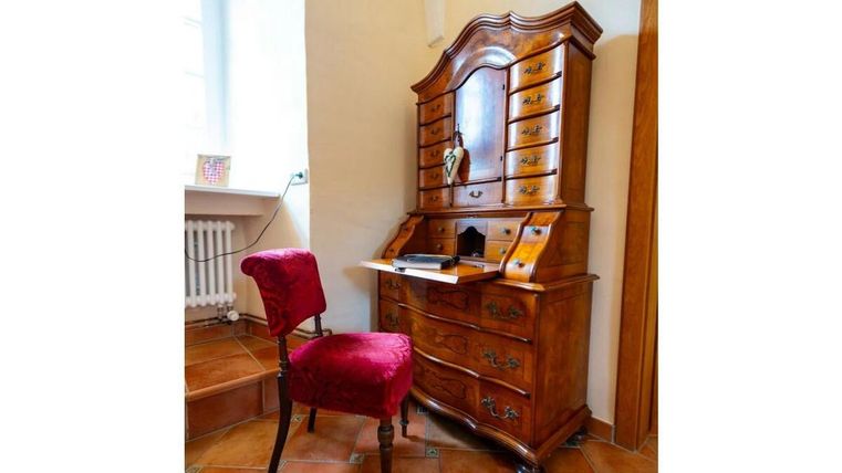Ein antikes Holzschreibtisch mit vielen Schubladen steht neben einem roten Stuhl. Der Raum hat helle Wände und einen gefliesten Boden.