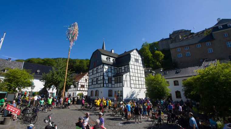 Ein belebter Platz mit vielen Radfahrern und einer festlichen Atmosphäre. Im Hintergrund sind historische Gebäude und Bäume zu sehen.